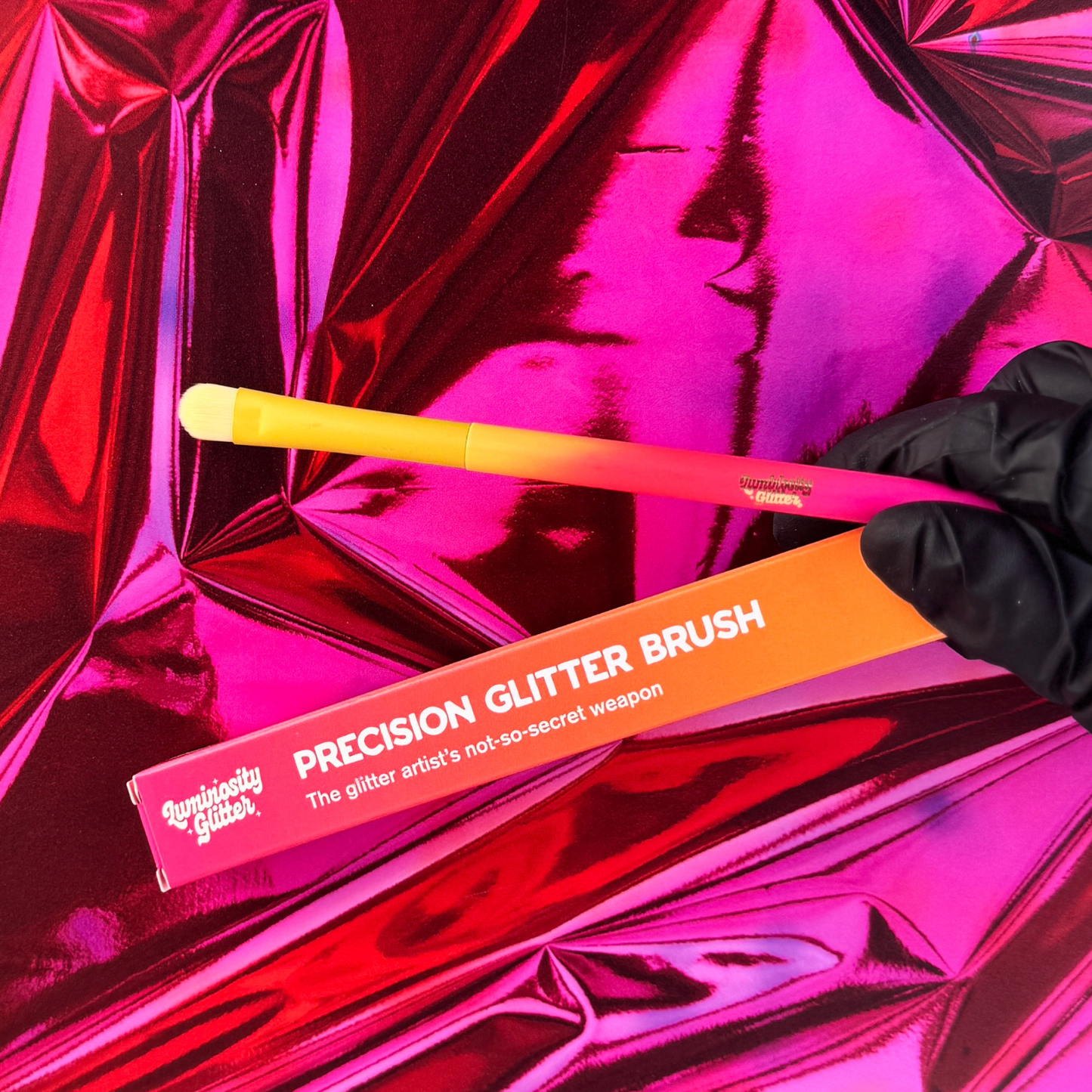 Precision glitter brush with orange handle and packaging on pink reflective background