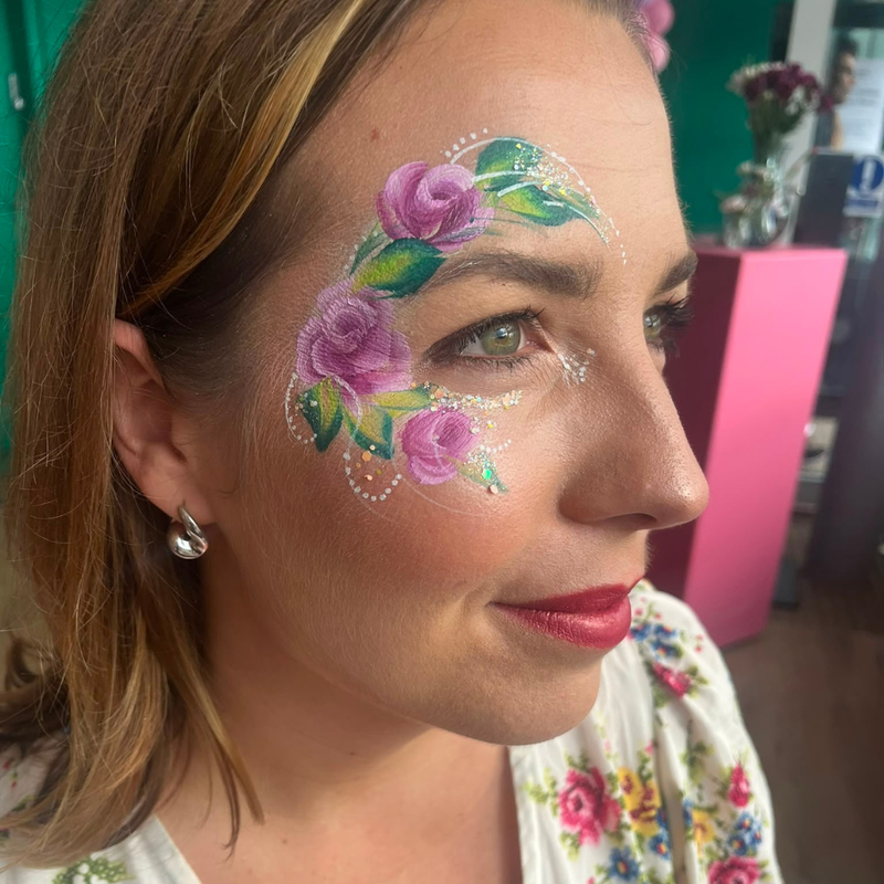 A close-up side profile of a woman with colorful floral face paint on her cheek, wearing a white top with a pink and green floral print, in what appears to be a salon or studio setting.