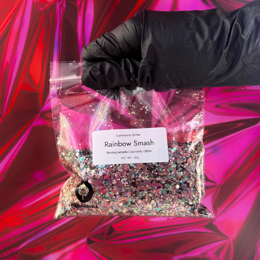 Bag of 'Rainbow Smash' glitter with a gloved hand holding it against a pink and red abstract background.