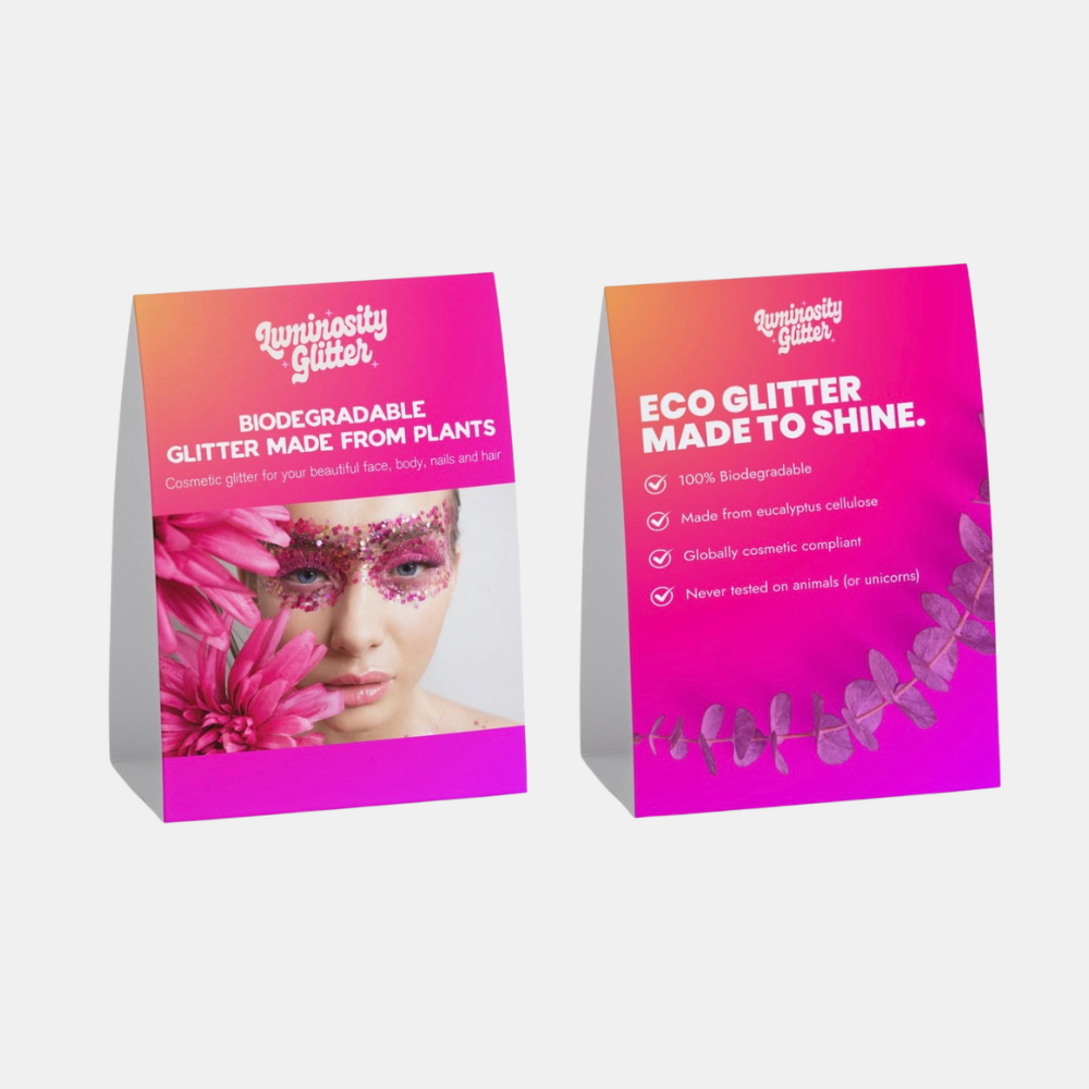 Two freestanding triangular product cards for Luminosity Glitter on a light gray background. The cards feature a bright pink-to-orange gradient design with white text and botanical graphics, and one card includes a model wearing glittery pink eye makeup.