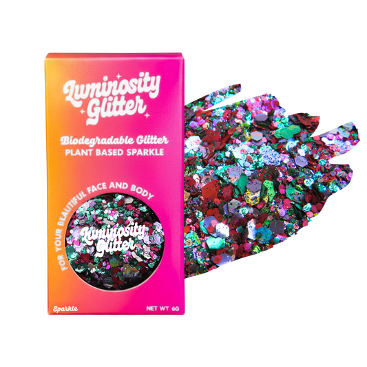 Luminosity Glitter packaging box in a vibrant pink-to-orange gradient, shown next to a hand displaying a mix of colorful, reflective biodegradable glitter flakes on a plain white background.