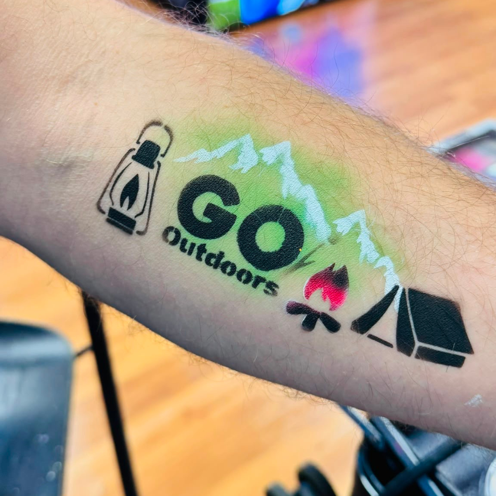 A temporary tattoo applied to a person's forearm, featuring a black lantern, the words 'GO Outdoors' in black, white mountain silhouettes, a small red and black campfire, and a black tent. The background shows a blurred indoor setting with wooden flooring.