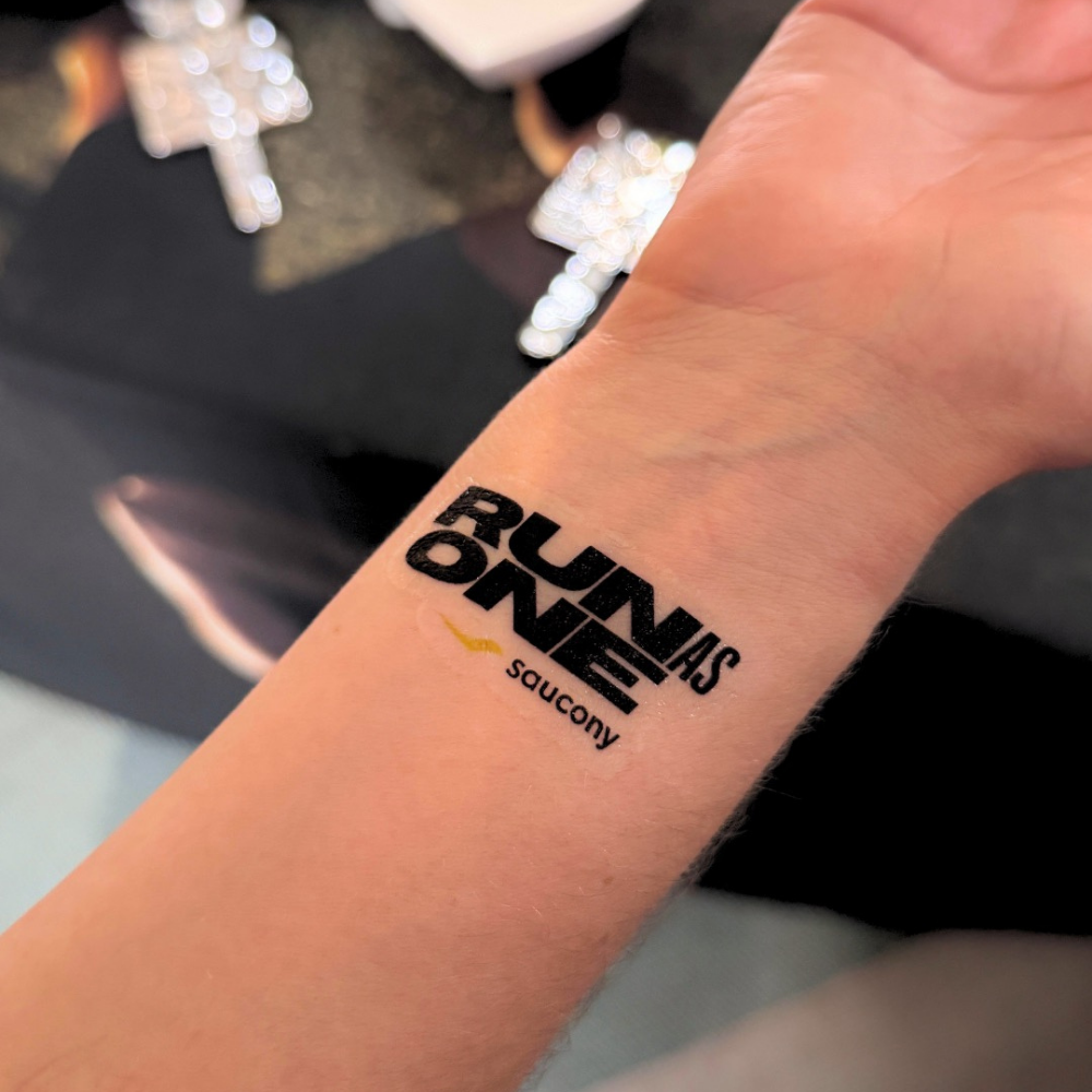 A close-up of a person's inner forearm showing a temporary tattoo with the text 'RUN AS ONE' and the 'saucony' logo in black and yellow.