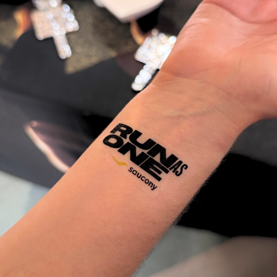 A close-up of a person's inner forearm showing a temporary tattoo with the text 'RUN AS ONE' and the 'saucony' logo in black and yellow.