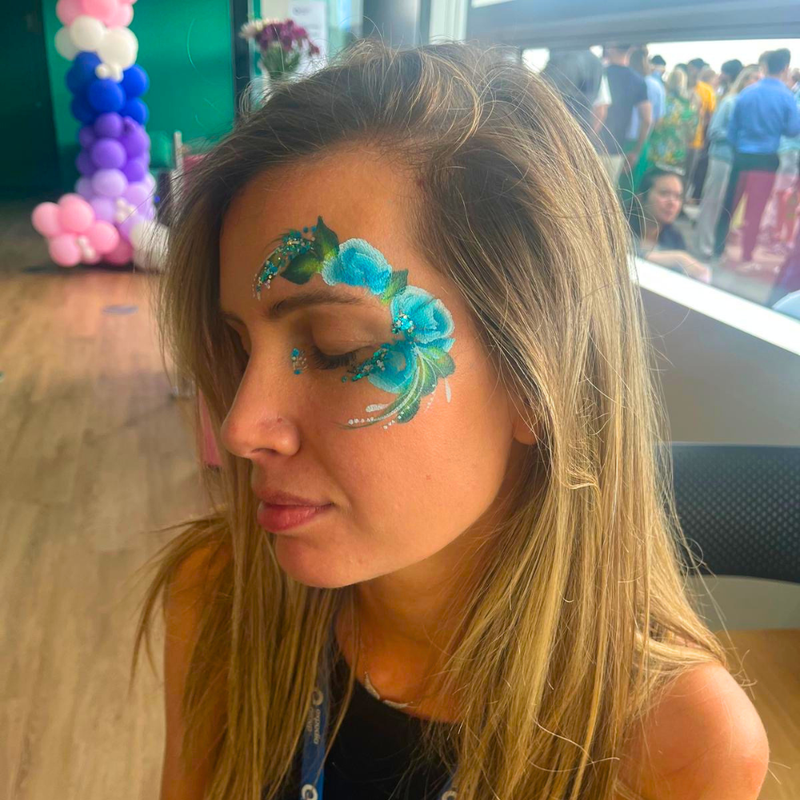 A woman with long blonde hair is shown in profile, wearing decorative face paint featuring blue flowers and green leaves on her forehead and temple. The background shows a crowded indoor event space with balloons and people.