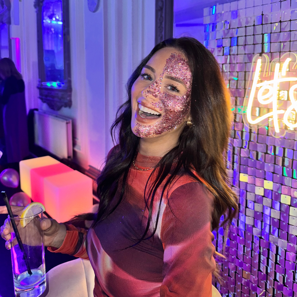 A beautiful woman with pink glitter on half of her face, smiling at a corporate event in London.