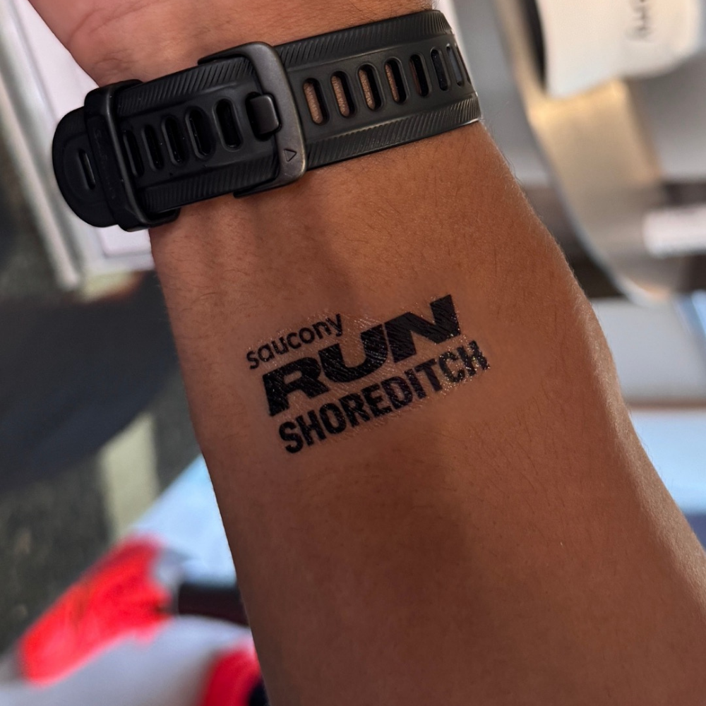 A close-up of a person's forearm wearing a black watch and featuring a black temporary tattoo that reads 'soucony RUN SHOREDITCH'.