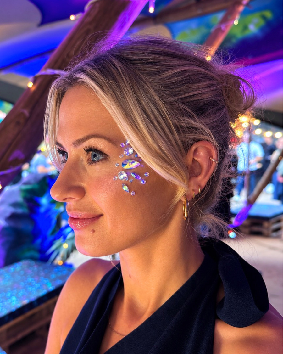 A woman with blonde hair in a bun wearing a black halter top, decorated with blue and purple rhinestone face gems, in a dimly lit venue with purple ambient lighting.