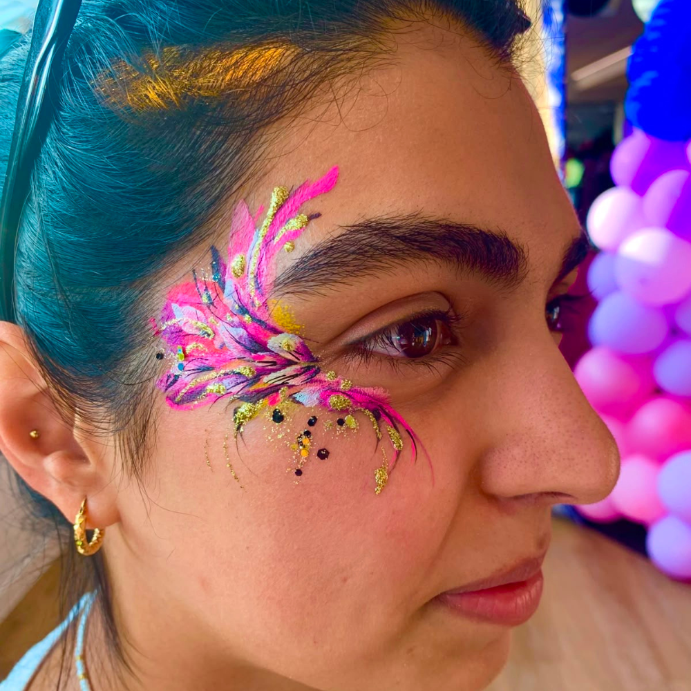 A close-up side profile of a woman with vibrant, colorful face paint on her cheek in bright pink, yellow, and blue, featuring small black and gold glitter dots. She has teal hair pulled back and is wearing a small gold hoop earring.
