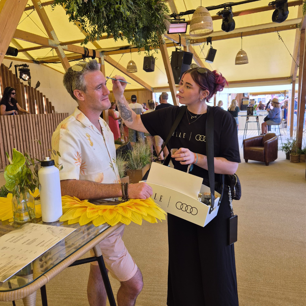 A woman in a black dress with 'ELLE & AUDI' printed on the front applies makeup to a man wearing a white patterned short-sleeve shirt and beige shorts at an outdoor event under a tent.