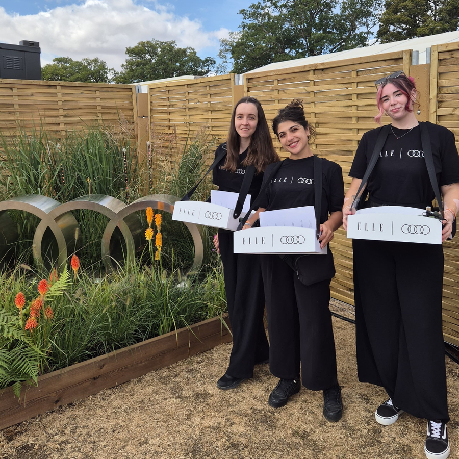 Three women standing outdoors in front of a wooden slat fence, each holding a white box printed with the brand name 'ELLE' and the Audi four-ring logo. They are dressed in matching black outfits.