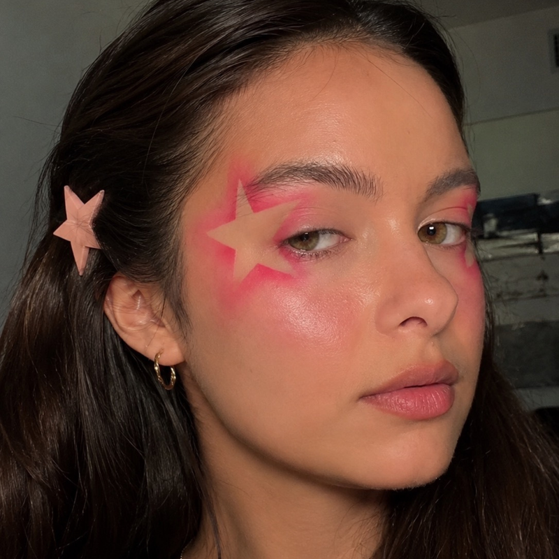 A close-up of a woman with pink star-shaped face makeup on her cheek and forehead, wearing a pink star hair clip in her dark hair.
