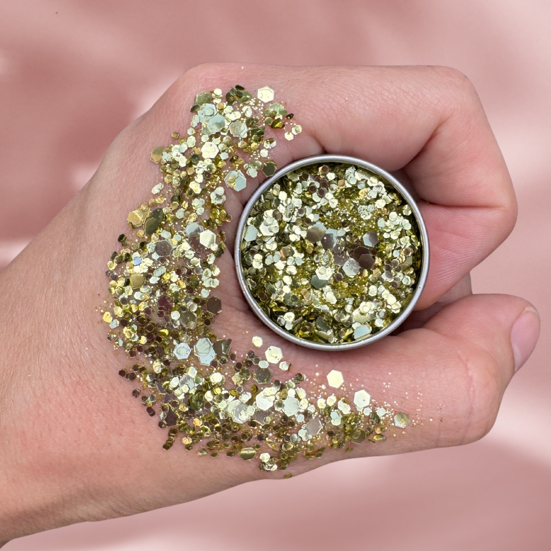 Hand holding a small container of gold glitter with more glitter on the palm against a pink background