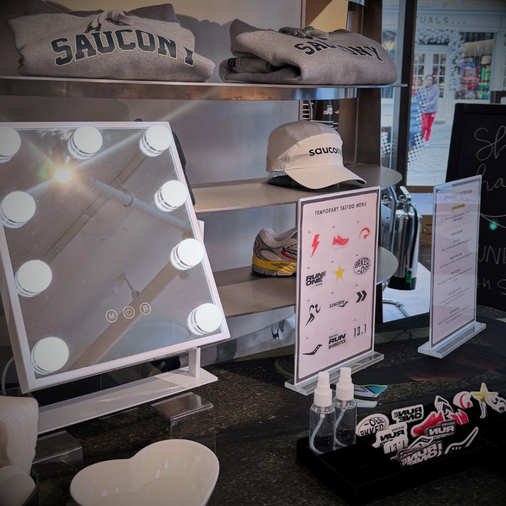 A retail display featuring a white vanity mirror with built-in light bulbs, small bottles, temporary tattoo stickers, and branded Saucony apparel including folded gray sweatshirts and a beige cap on shelves.